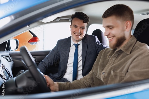 A joyful car dealership employee shows new car options to a male buyer sitting in the salon.