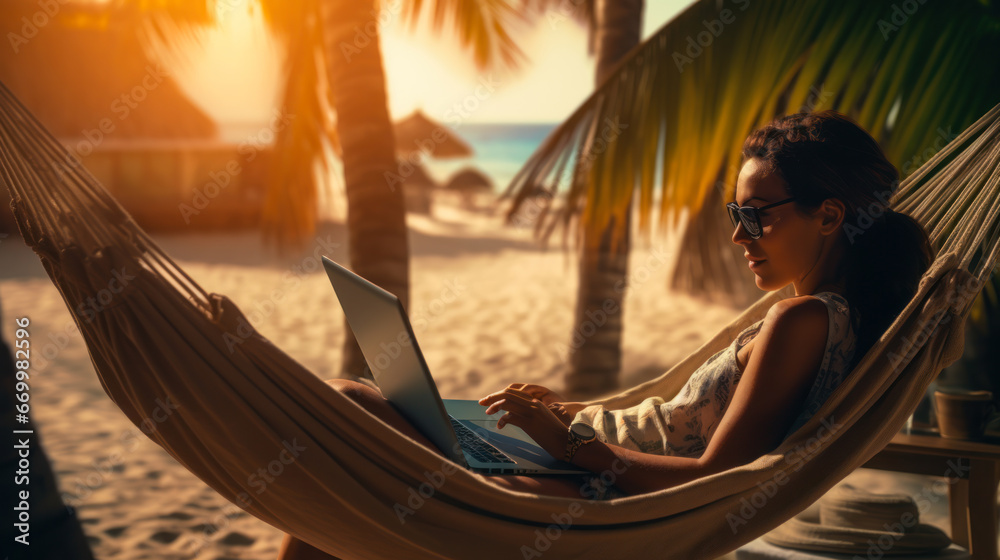 Young freelancer woman using a laptop while lying in a hammock on a sandy beach under palm trees on the seashore. Concept of remote work, vacation, relaxation.
