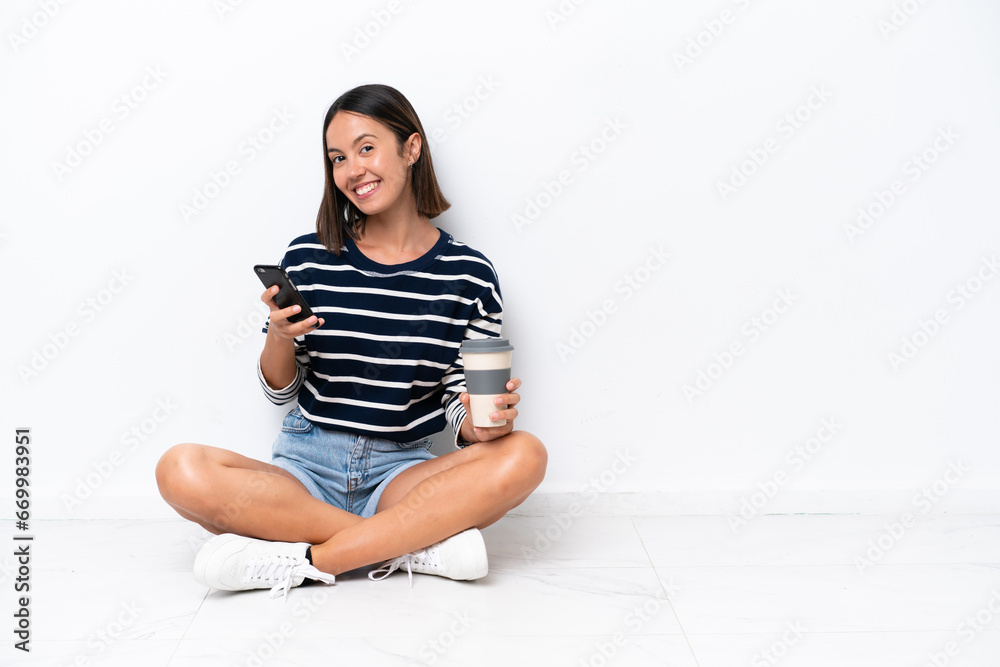 Young caucasian woman sitting on the floor isolated on white background holding coffee to take away and a mobile Young caucasian woman sitting on the floor isolated on white background holding coffee to take away and a mobile
