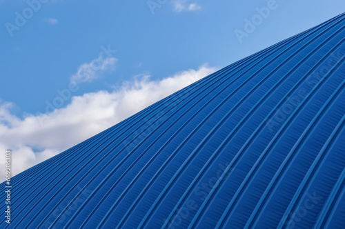 A fragment of a hangar made of blue rolled metal against the background of the sky with clouds. A simple metal structure of an industrial building from the outside.