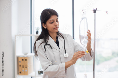 Adult female doctor in white lab coat standing near medical dropper and controlling liquid medication. Indian woman preparing infusion drip for intravenous treatment system.