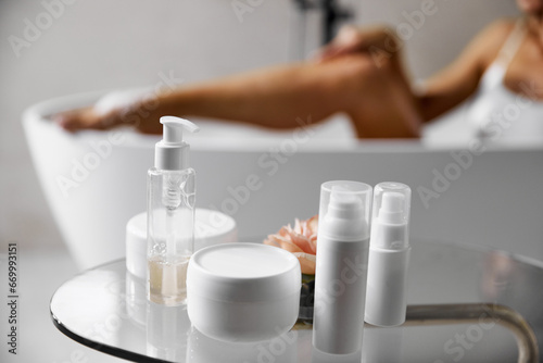 Morning time. Table top with cosmetic treatment  skincare products and blur bathroom with lying bathtub young pretty woman with slim  fit legs.