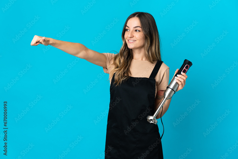 Romanian woman using hand blender isolated on blue background giving a thumbs up gesture Romanian woman using hand blender isolated on blue background giving a thumbs up gesture