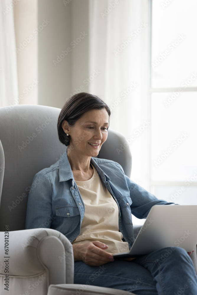 Interested middle-aged woman read news on laptop, spend time on internet settled down on armchair at home. Modern wireless tech usage for business, gaining new skills, searching information on-line Interested middle-aged woman read news on laptop, spend time on internet settled down on armchair at home. Modern wireless tech usage for business, gaining new skills, searching information on-line