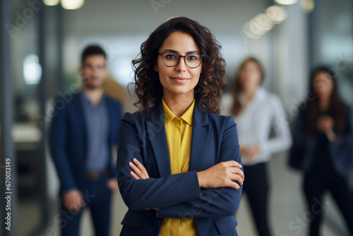 Portrait of one confident young hispanic business woman standing with arms crossed in an office with her colleagues in the background, Ambitious entrepreneur and determined leader ready for success  photo