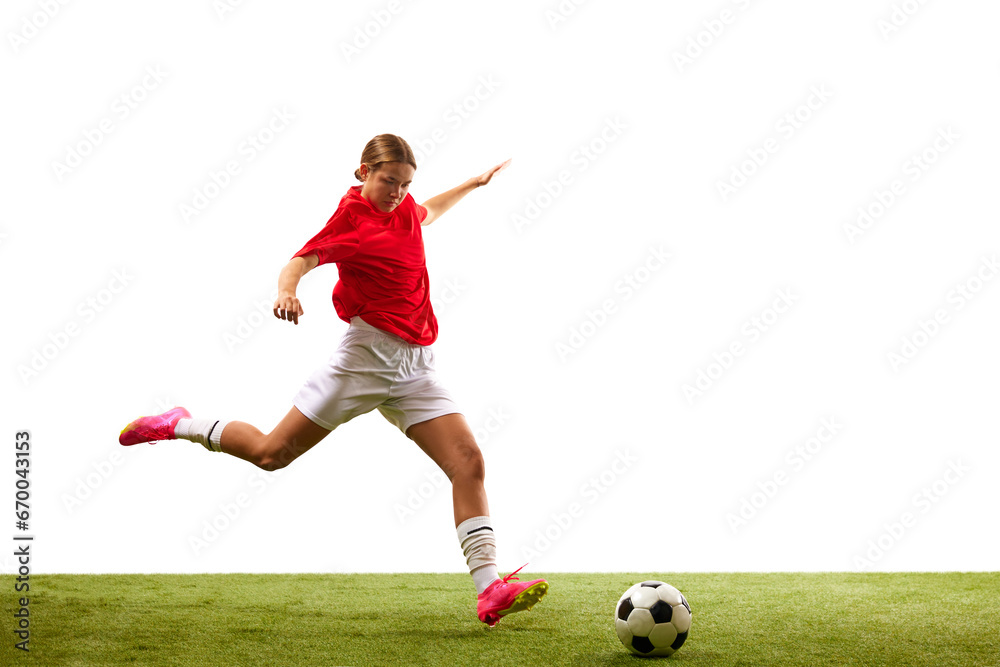 Young concentrated girl, football player in motion, playing on green grass, ready to hit ball isolated on white background. Concept of sport, competition, action, success, winner. Copy space for ad