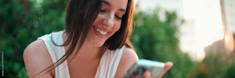 Cute young woman with freckles and dark loose hair wearing white top is using mobile phone. Beautiful girl sits in city park looking through photos, videos on mobile phone. Camera moves around Cute young woman with freckles and dark loose hair wearing white top is using mobile phone. Beautiful girl sits in city park looking through photos, videos on mobile phone. Camera moves around