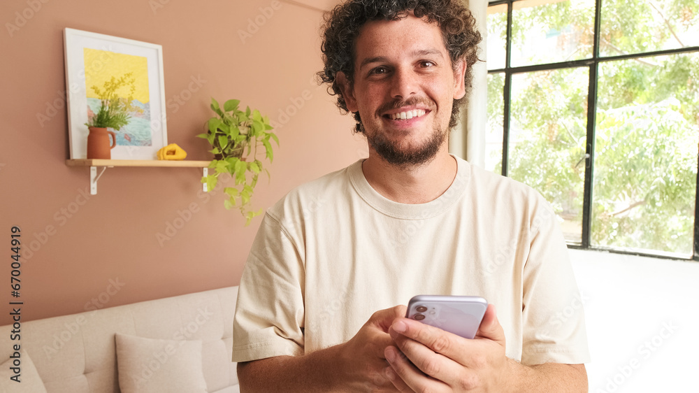 Smiling young man with curly hair and beard using mobile phone while standing in cozy living room Smiling young man with curly hair and beard using mobile phone while standing in cozy living room