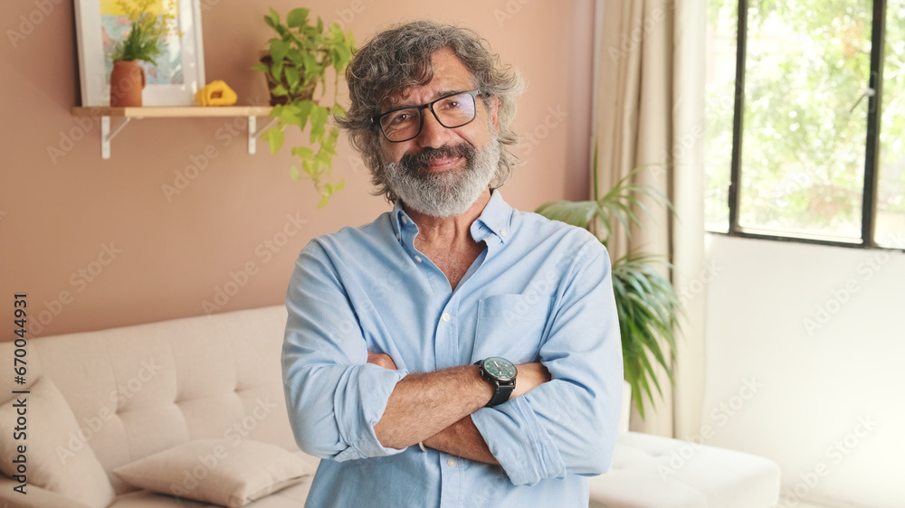 An elderly man crosses his arms and looks at the camera with smile in cozy living-room