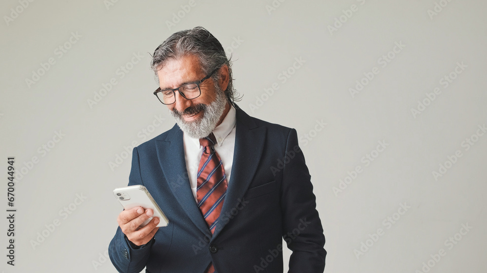 Businessman in glasses with gray hair, looking with smile at the screen of mobile phone, isolated on gray background in the studio Businessman in glasses with gray hair, looking with smile at the screen of mobile phone, isolated on gray background in the studio