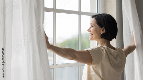 Side view rested middle-aged woman standing near window, open curtains, admires city view, enjoy peaceful, quiet morning, woke up early on weekend, welcoming, start new day, full of hope of happy day