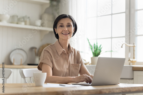 Attractive mature woman use laptop sit at table in kitchen, typing on device, do telework from home, smile look at camera. Modern tech usage for leisure, work, on-line electronic services, e-bank app