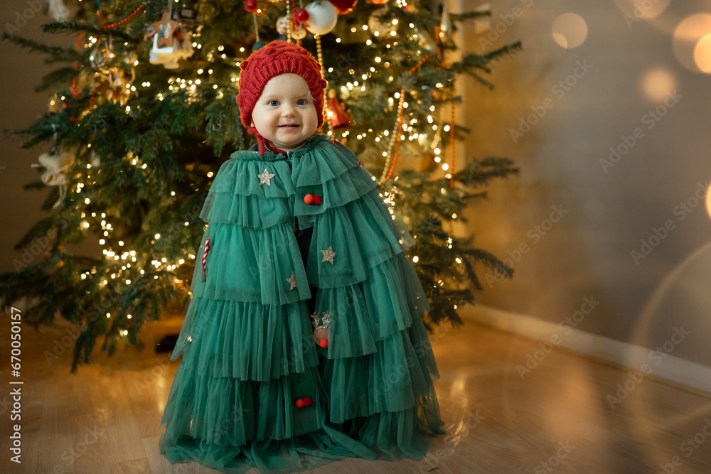 Smiling Little baby girl in a Christmas tree carnival costume and warm winter hat stands in front of a Christmas tree . New Year celebration . High quality photo