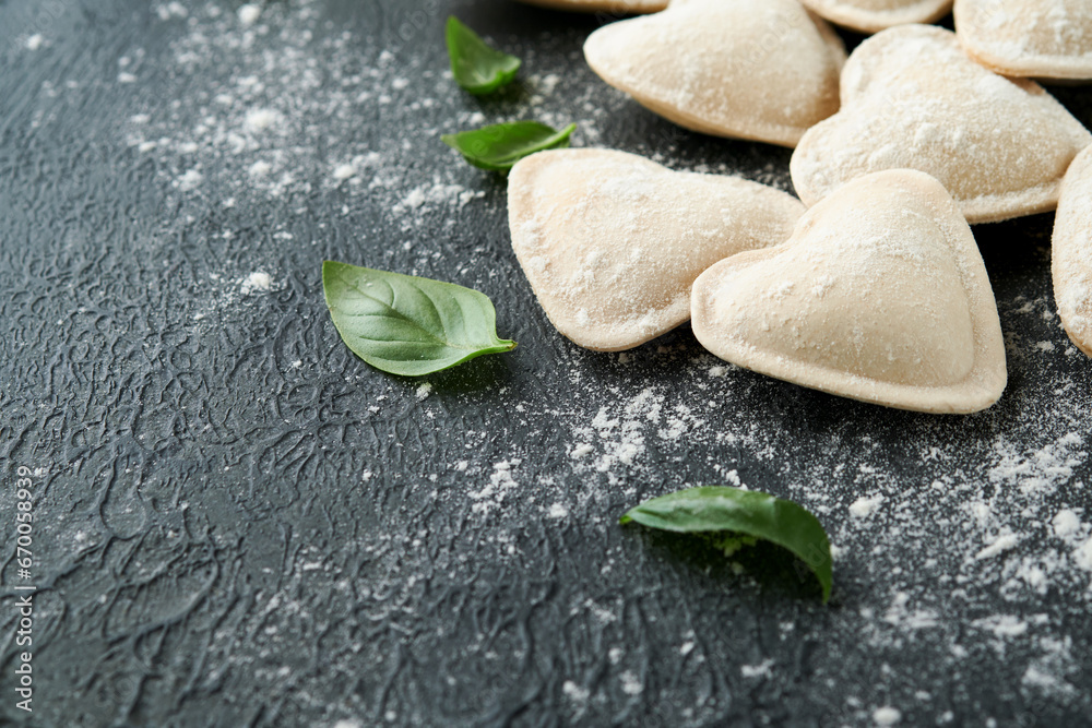 Fototapeta premium Italian ravioli pasta in heart shape. Tasty raw ravioli with flour and basil on dark background. Food cooking ingredients background. Valentines or Mothers Day lunch ideas. Top view with copy space.