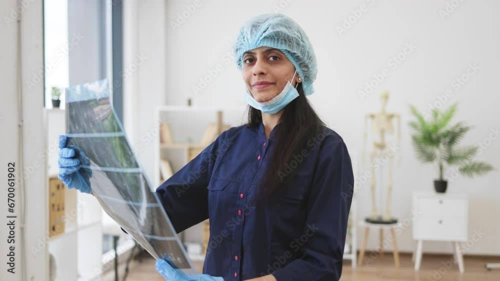 Portrait of hindu medical worker dressed in dark blue uniform and disposable cap holding result of x-ray scan of sick patient in cabinet. Competent doctor investigating diseases before surgery. Portrait of hindu medical worker dressed in dark blue uniform and disposable cap holding result of x-ray scan of sick patient in cabinet. Competent doctor investigating diseases before surgery.