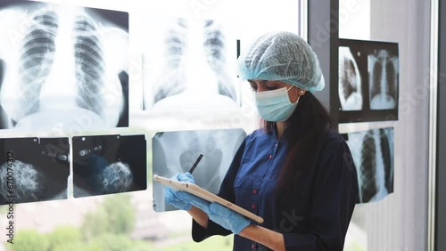 Focused female medical worker in face mask and scrub top posing with digital tablet near x-ray scans fixed on window glass. Qualified hindu surgeon in medical gloves studying injuries. photo