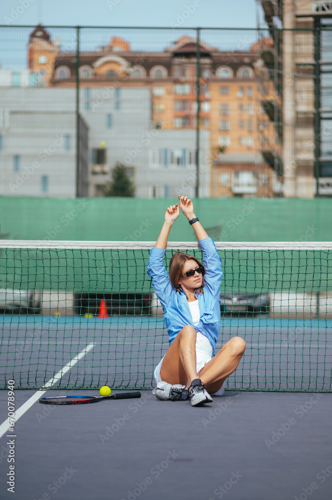 Attractive female tennis player in shirt sitting on court near net with arms raised and posing for camera