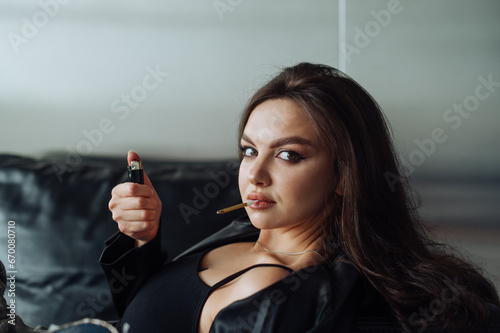 Quadro em tela Close-up portrait of an attractive woman in dark clothes lying on a leather sofa with a candle in her mouth and a lighter in her hand posing for the camera with a serious face