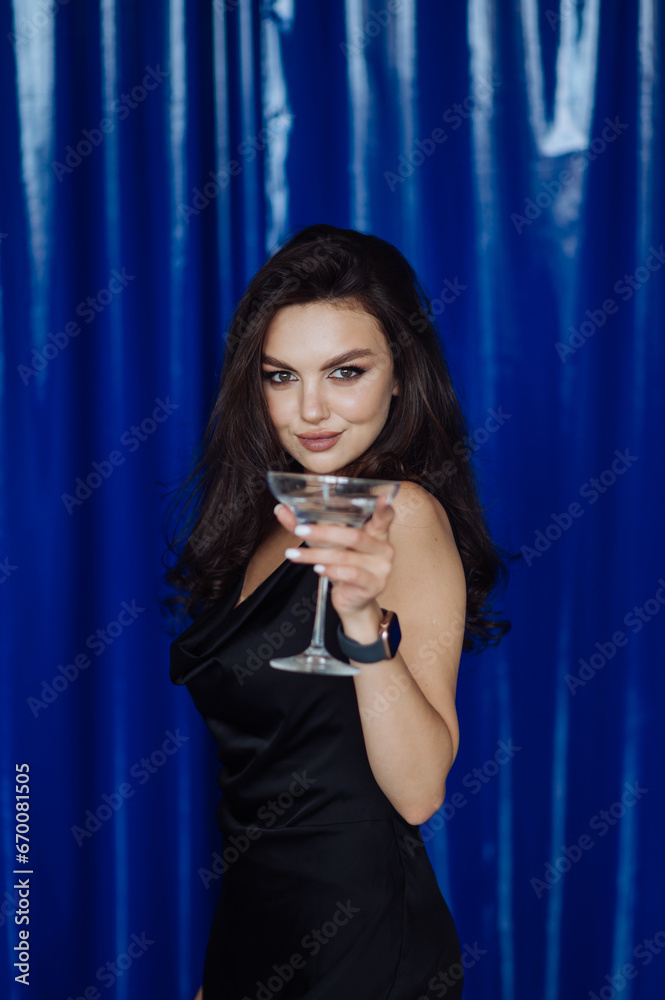 An attractive brunette with a glass of alcohol in her hand stands on a blue background and looks at the camera with a smile on her face. An attractive brunette with a glass of alcohol in her hand stands on a blue background and looks at the camera with a smile on her face.