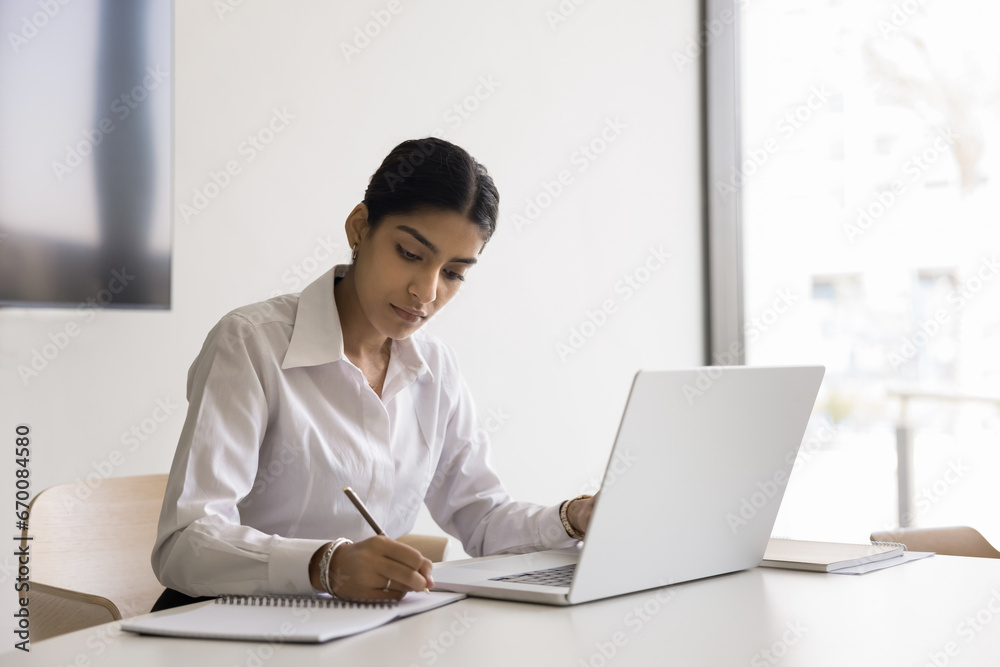 Serious pretty Indian business professional girl writing notes at laptop compute, sitting at workplace table in office, talking to client on video conference call, studying online Serious pretty Indian business professional girl writing notes at laptop compute, sitting at workplace table in office, talking to client on video conference call, studying online