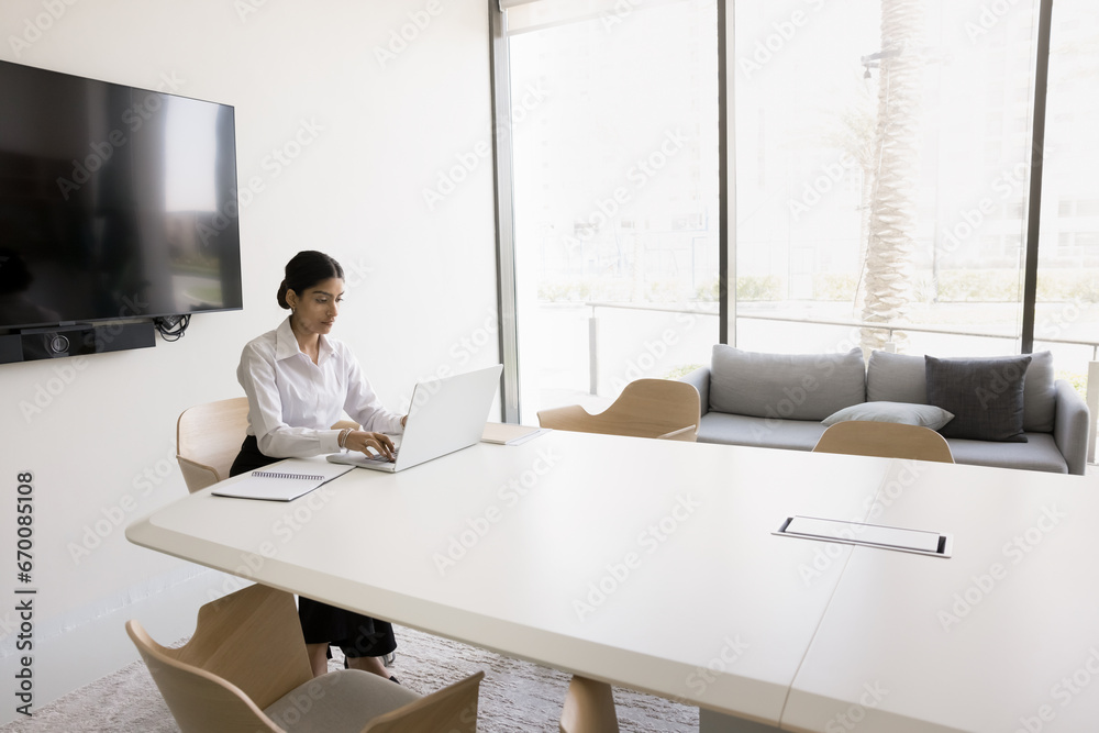 Confident young Indian business professional woman working at laptop alone in meeting room, sitting at large table in boardroom, typing on computer, using Internet communication for job
