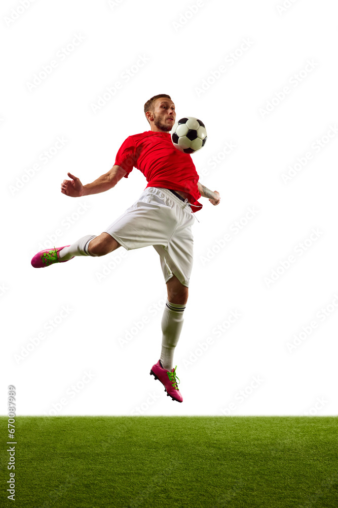 Young man, professional soccer player in red sportwear and boots performing football tricks against white background with green grass.