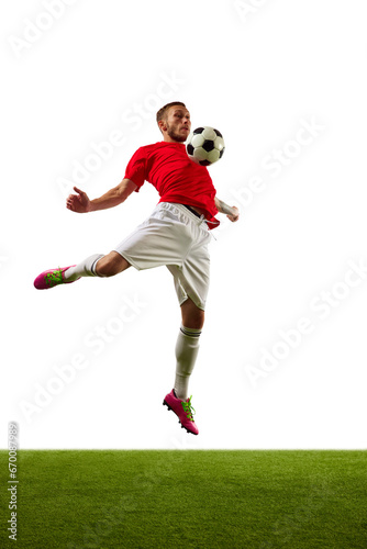 Young man, professional soccer player in red sportwear and boots performing football tricks against white background with green grass. © Lustre