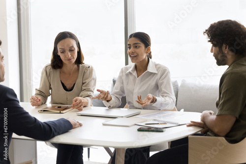 Positive motivated young Indian professional woman offering creative idea for project to listening coworkers on brainstorming meeting, sitting at table, speaking, smiling, working with team