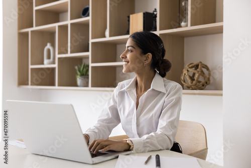 Cheerful successful young Indian manager woman using laptop in office, looking away, thinking on creative ideas for business project, enjoying professional inspiration, job success photo