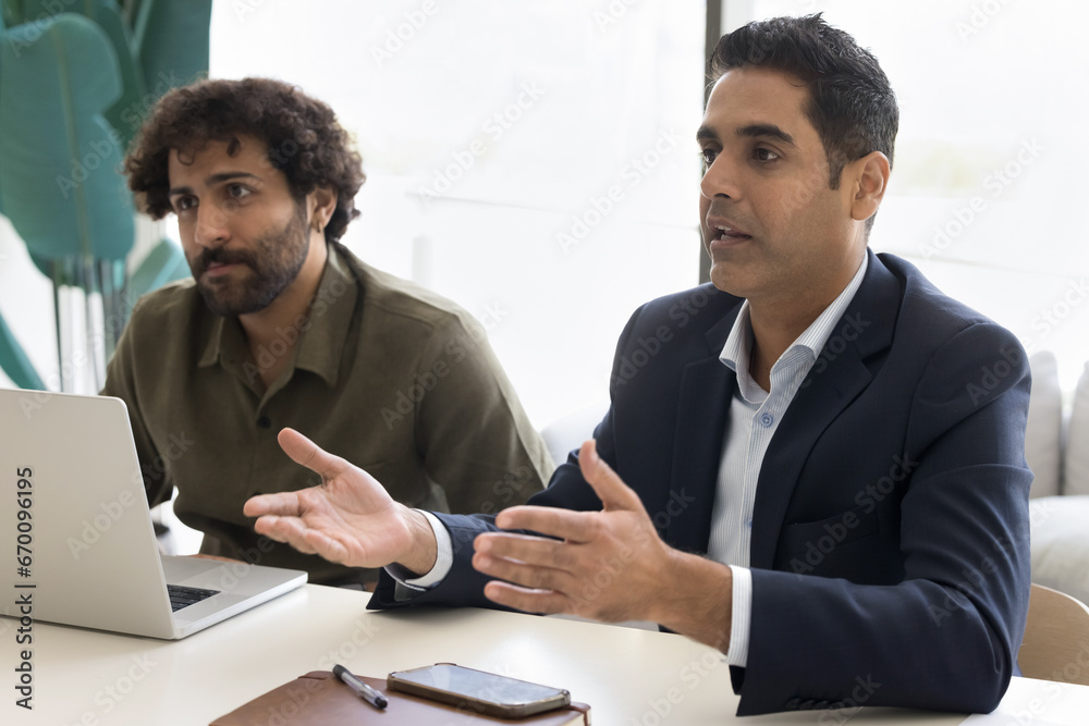 Serious Indian businessman in formal jacket talking to partners or colleagues on negotiation meeting at table. Diverse business colleagues discussing cooperation, project management, teamwork Serious Indian businessman in formal jacket talking to partners or colleagues on negotiation meeting at table. Diverse business colleagues discussing cooperation, project management, teamwork
