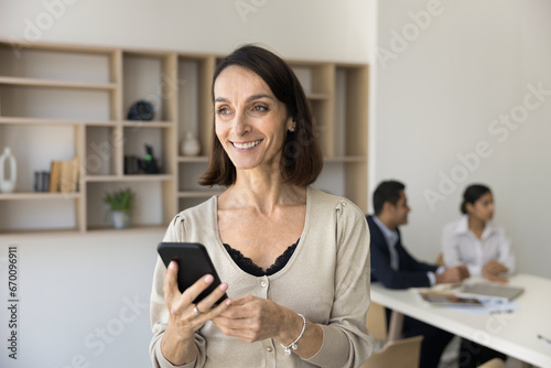 Happy middle aged business professional woman using work online technology on mobile phone, looking away, thinking on successful project, smiling, laughing while tam talking in blurred background