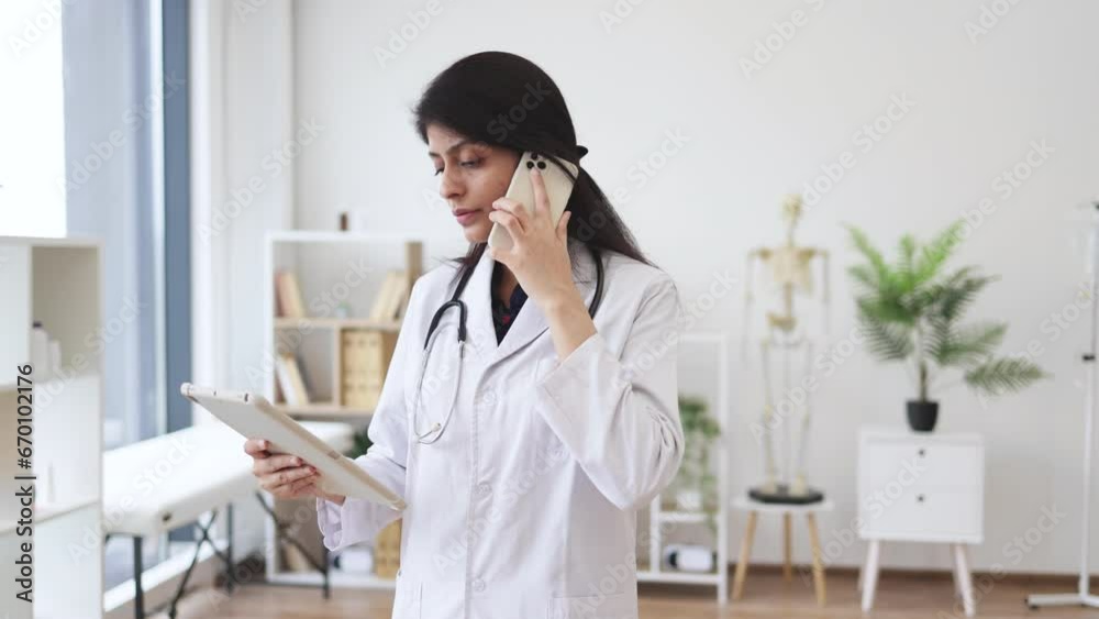 Front view of female physician standing and talking on mobile phone while looking on tablet screen. Competent hindu doctor using modern devices for work and consultation from hospital. Front view of female physician standing and talking on mobile phone while looking on tablet screen. Competent hindu doctor using modern devices for work and consultation from hospital.