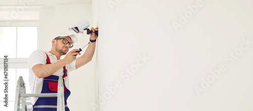 Male technician adjusts, installs or repairs modern CCTV camera on white wall in office or home. Man with screwdriver on ladder adjusting smart CCTV surveillance system. Copy space. Web banner.