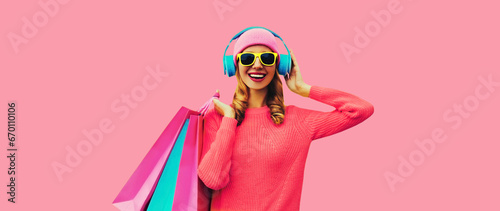 Portrait of stylish happy smiling young woman with colorful shopping bags posing listening to music in headphones wearing knitted sweater, hat on pink studio background