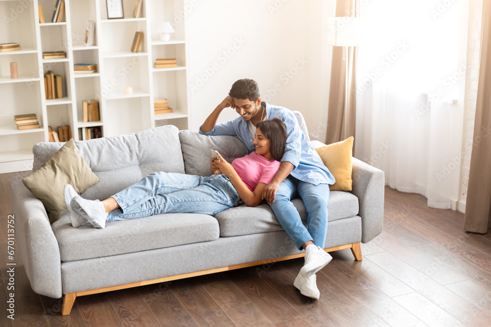 Young Indian couple enjoying leisure time on sofa with phone Young Indian couple enjoying leisure time on sofa with phone