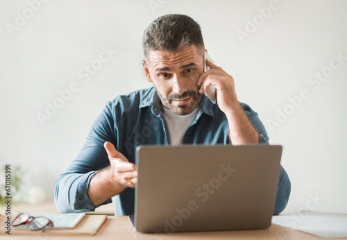 Displeased Man Talking On Cellphone Gesturing To Laptop At Workplace