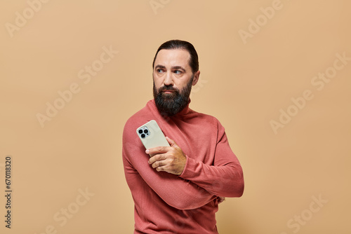 portrait of handsome bearded man in turtleneck jumper holding smartphone on beige background