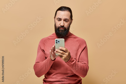 portrait of handsome bearded man in turtleneck jumper using smartphone on beige background