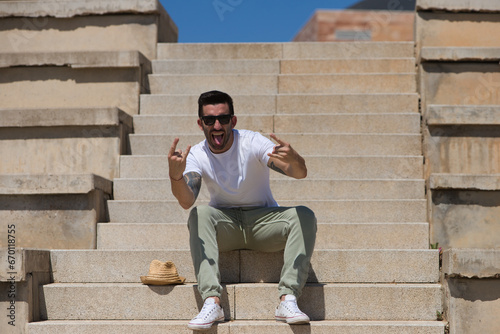Fotografía Handsome young man dressed in casual clothes and sunglasses sitting on the steps of the park making a horn gesture with his two hands