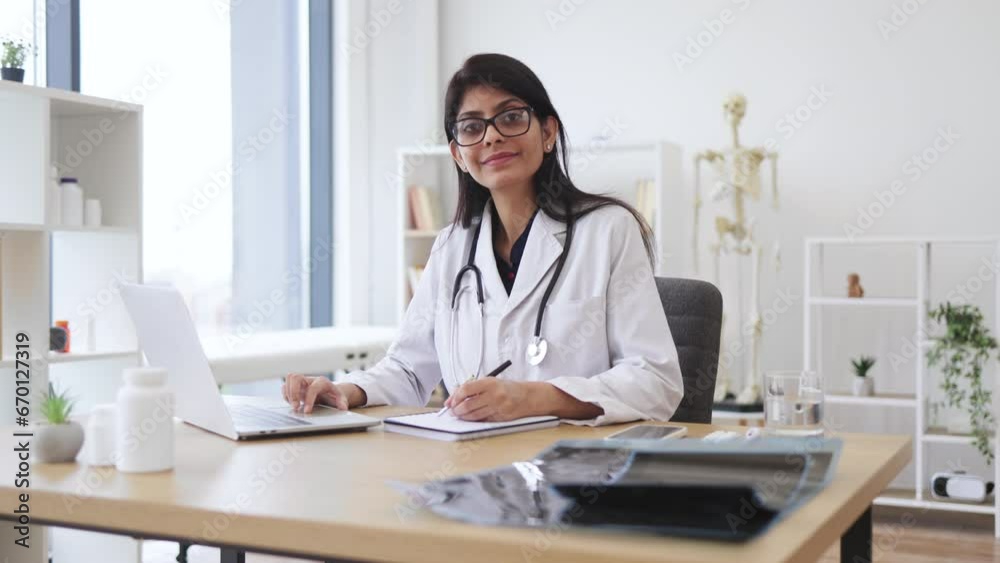 Side view of indian female doctor using portable computer while sitting at office desk in health center interior. Efficient mature general practitioner checking patient records via gadget. Side view of indian female doctor using portable computer while sitting at office desk in health center interior. Efficient mature general practitioner checking patient records via gadget.