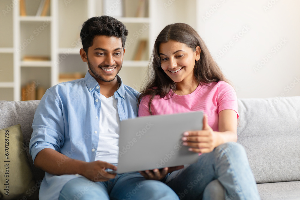 Cheerful young Indian couple on couch, sharing a laptop moment Cheerful young Indian couple on couch, sharing a laptop moment