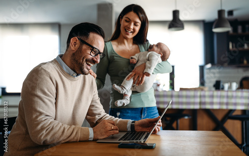 Young father works on laptop while his wife looks after child.Work from home parenting telecommuting