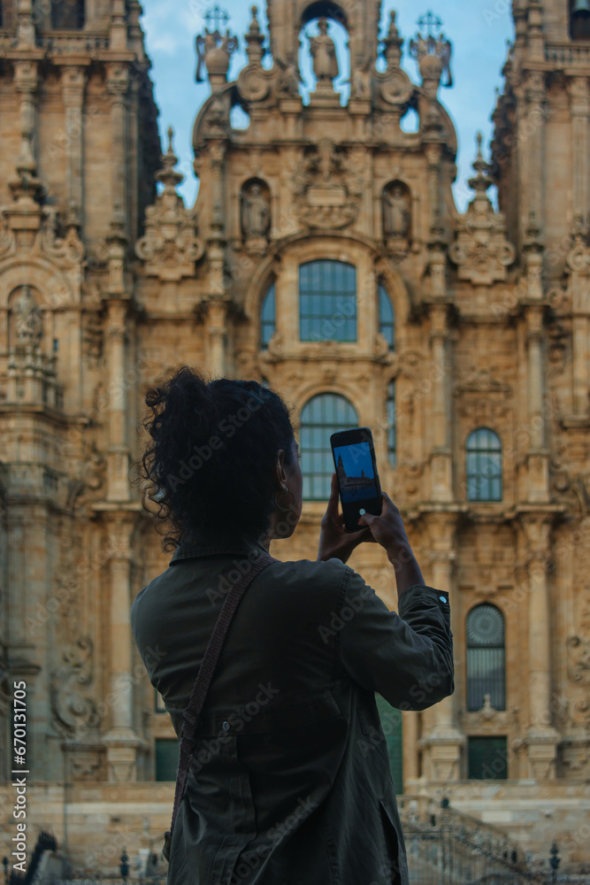 Young Hispanic woman with curly hair taking a photograph with her mobile phone of the facade of the Cathedral of Santiago de Compostela Young Hispanic woman with curly hair taking a photograph with her mobile phone of the facade of the Cathedral of Santiago de Compostela