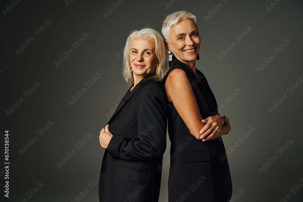 happy senior female models in black classic attire posing back to back and smiling at camera on grey