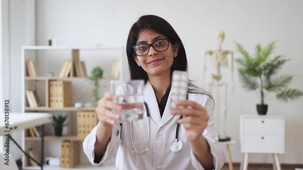 Front view of pleasant female doctor sitting at table with modern gadgets while holding glass of water and pills in hands. Smiling indian specialist showing method of treating disease at clinic. Front view of pleasant female doctor sitting at table with modern gadgets while holding glass of water and pills in hands. Smiling indian specialist showing method of treating disease at clinic.