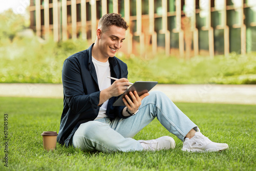 Happy millennial european man enjoy lifestyle, work on tablet with coffee cup takeaway, sit on grass in city