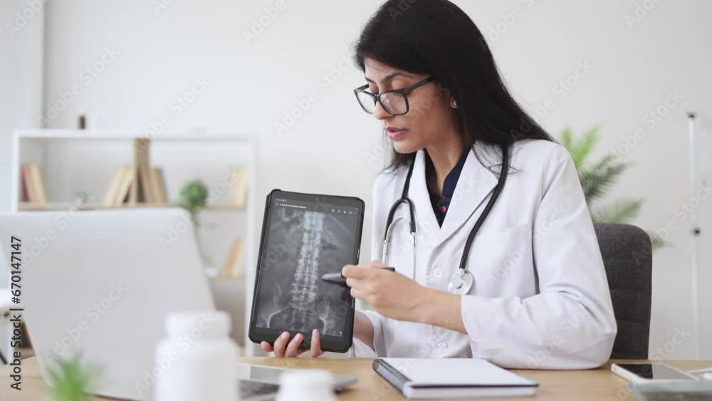 Mature female physician in white coat sitting at desk and pointing out at spine x-ray on digital tablet. Indian specialist demonstrating often found disease using modern gadget at cozy workplace. Mature female physician in white coat sitting at desk and pointing out at spine x-ray on digital tablet. Indian specialist demonstrating often found disease using modern gadget at cozy workplace.