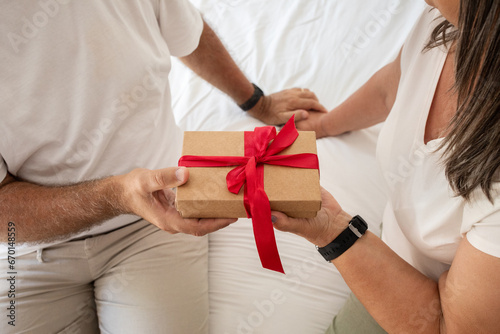Positive senior caucasian woman gives gift box to man, sit on bad in bedroom interior