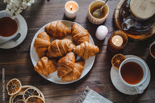 small croissants on a plate, a cozy hygge atmosphere, a glass teapot and cups of tea nearby. wooden background, lilac in a vase, top view