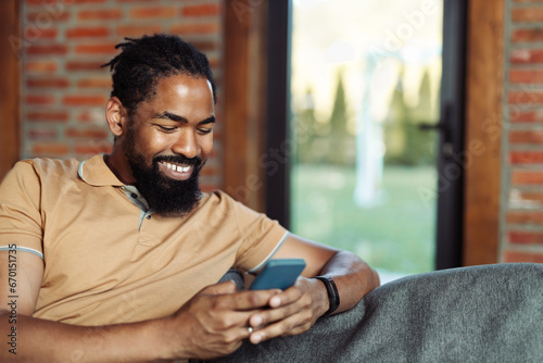 Happy African American man relaxing on sofa in the living room and reading a text message on his mobile phone